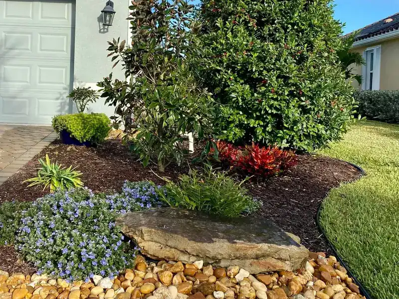 Garage-corner bed with blue daze, a flat boulder and a river rock border along a crisp lawn edge, by The Master’s Touch Landscape and Design