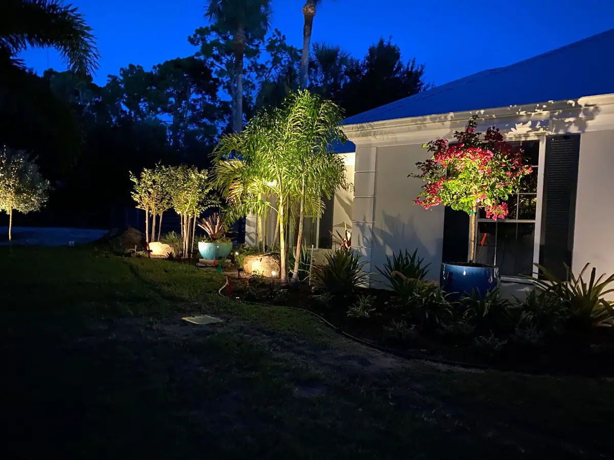 Uplit palms, pots and standards at dusk around a white home with a blue metal roof, by The Master’s Touch Landscape and Design