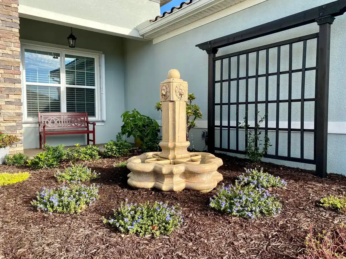 Pedestal fountain in a fresh mulch bed with blue daze, set against a black grid trellis, by The Master’s Touch Landscape and Design