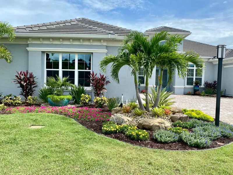 Blue-grey home with a triple palm, sandstone boulders, red ti plants and a curved steel-edged bed, by The Master’s Touch Landscape and Design