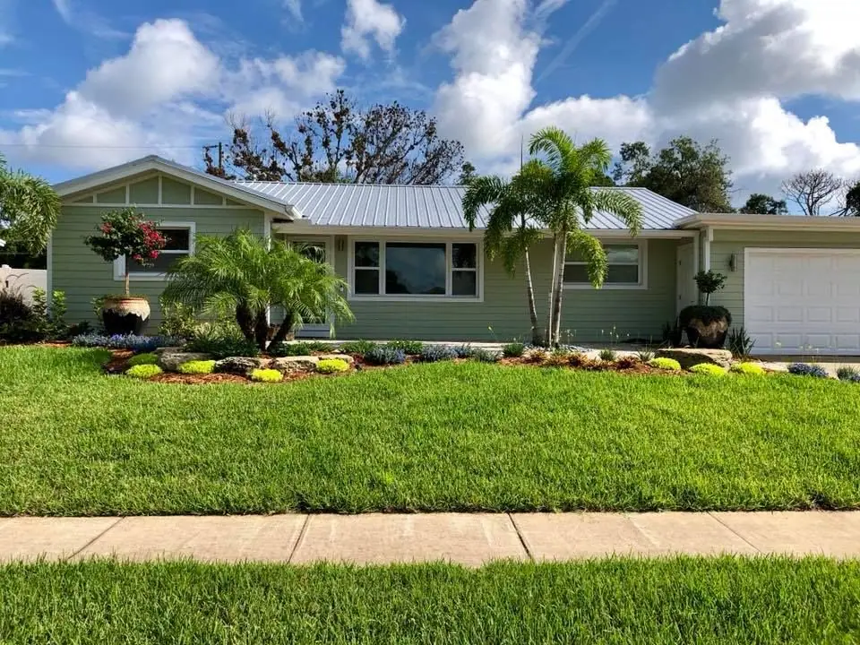 Sage green ranch home with a wide lawn and a long planted bed with palms, by The Master’s Touch Landscape and Design