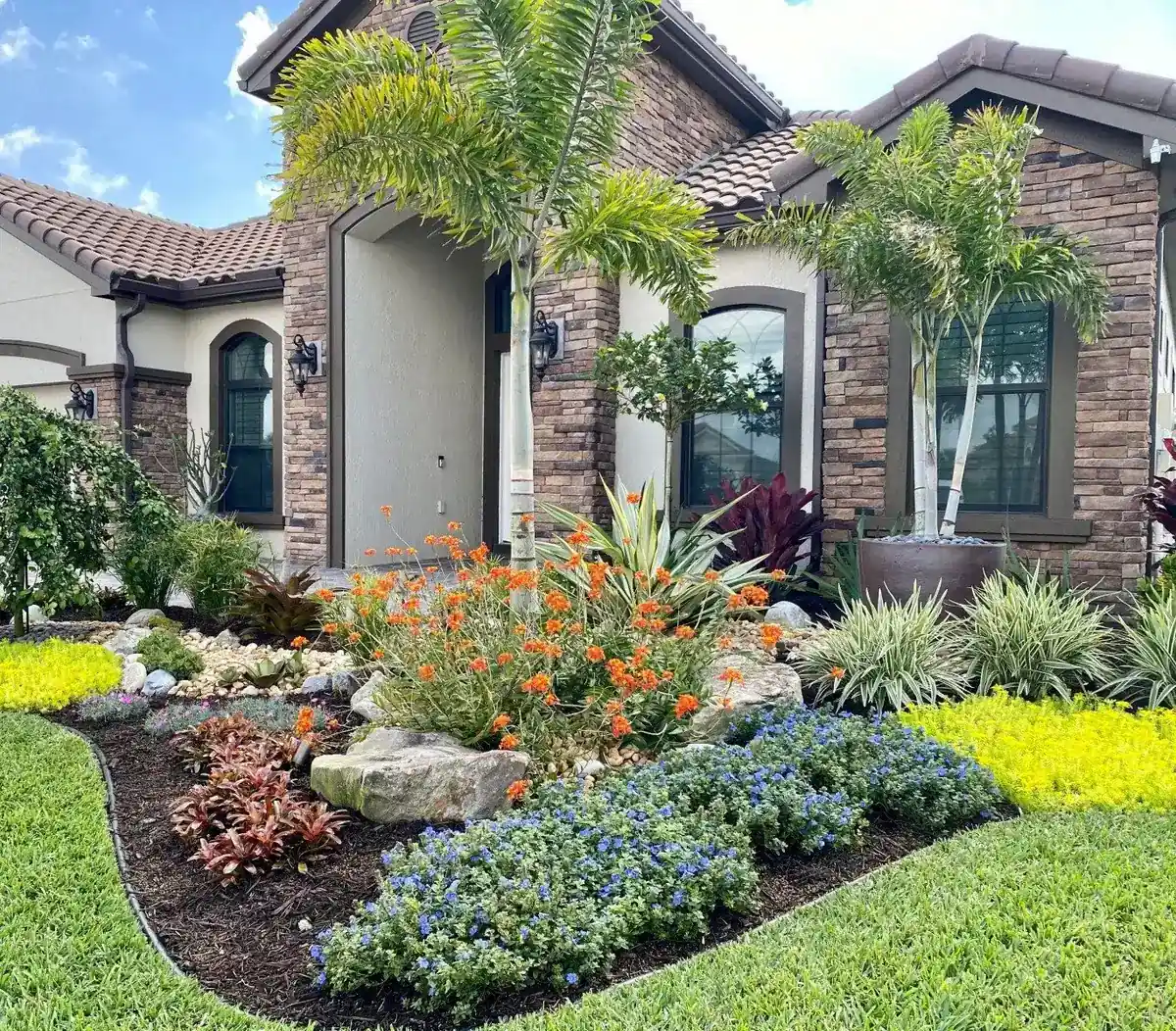 Stone-front home with a curved, steel-edged entry bed of foxtail palms, boulders, blue daze and golden groundcover, by The Master’s Touch Landscape and Design