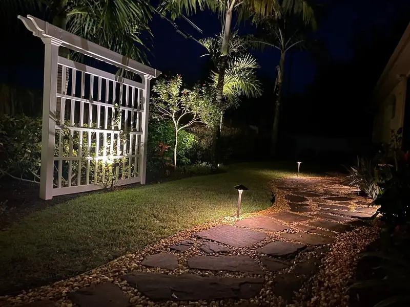 White lattice arbor and a flagstone path lit by low path lights at night, by The Master’s Touch Landscape and Design