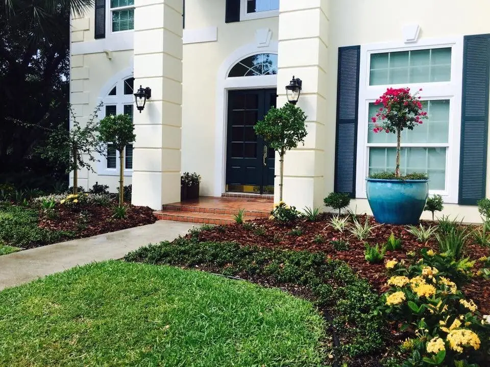 White two-storey home with dark shutters, a cobalt pot, topiary standards and dark mulch beds along a curved lawn, by The Master’s Touch Landscape and Design