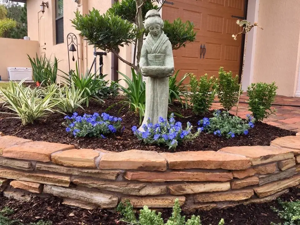 Curved stacked-stone garden wall holding a raised bed with blue flowers and a statue, in Melbourne, by The Master’s Touch Landscape and Design