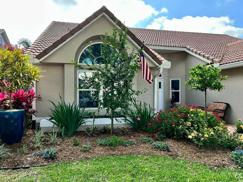 Tile-roofed home with new foundation beds, red ti plants in a blue pot and a young standard tree, by The Master’s Touch Landscape and Design