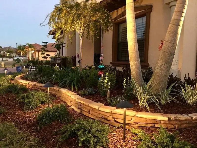 Path lights and uplights on a stacked-stone bed wall and palms at dusk, by The Master’s Touch Landscape and Design